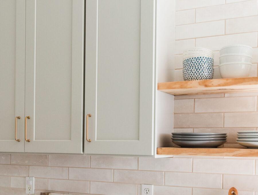 Light gray shaker style cabinets with gold handles, next to light wood floating shelves with plates and bowls