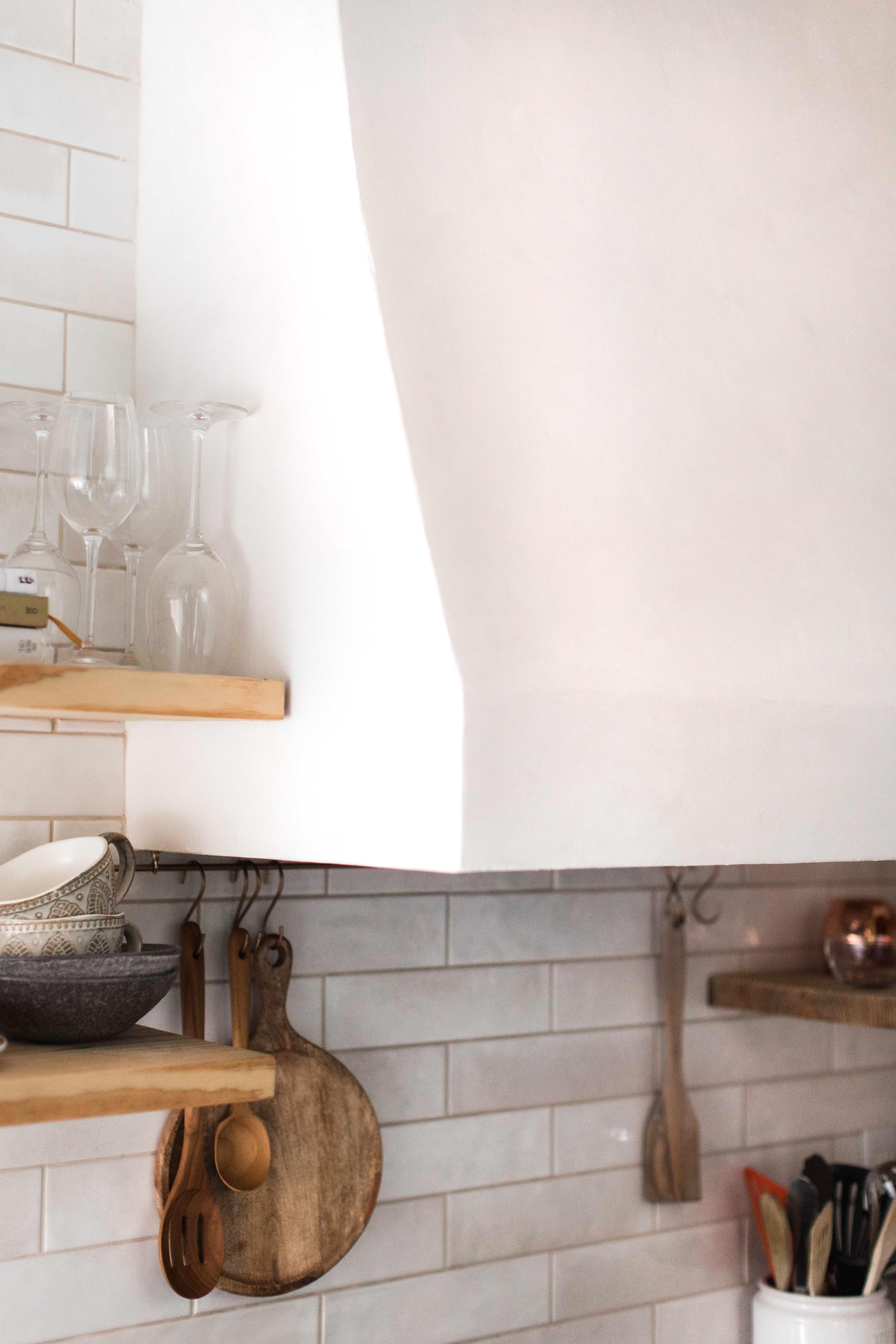 White venetian plaster range hood next to floating light wood shelves with kitchenware